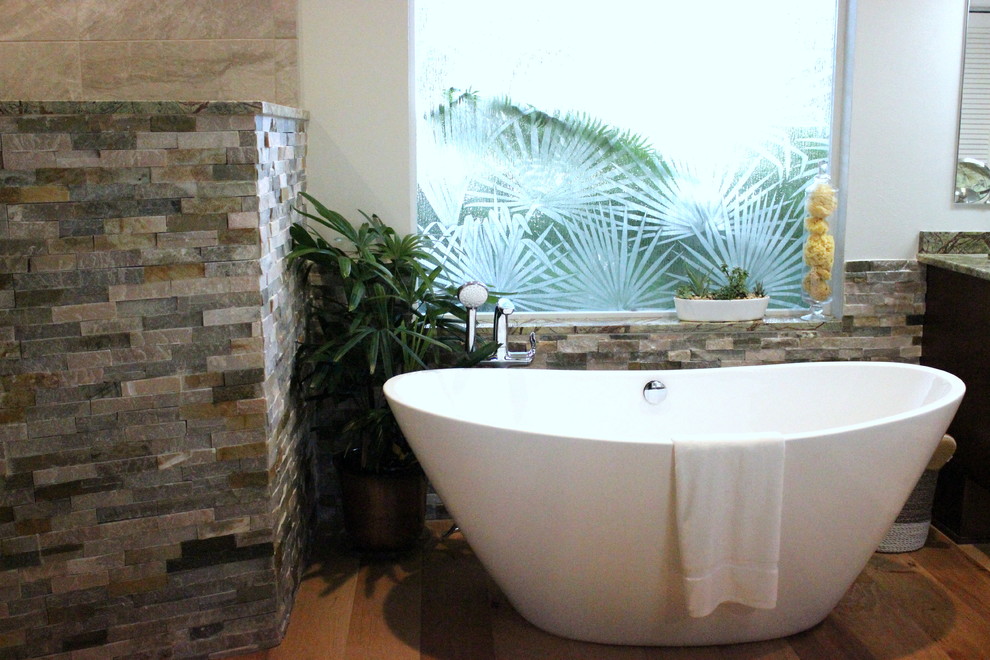 This is an example of a large tropical master bathroom in Tampa with flat-panel cabinets, dark wood cabinets, a freestanding tub, a corner shower, beige tile, brown tile, gray tile, multi-coloured tile, stone tile, grey walls, light hardwood floors, a vessel sink, granite benchtops and a two-piece toilet.