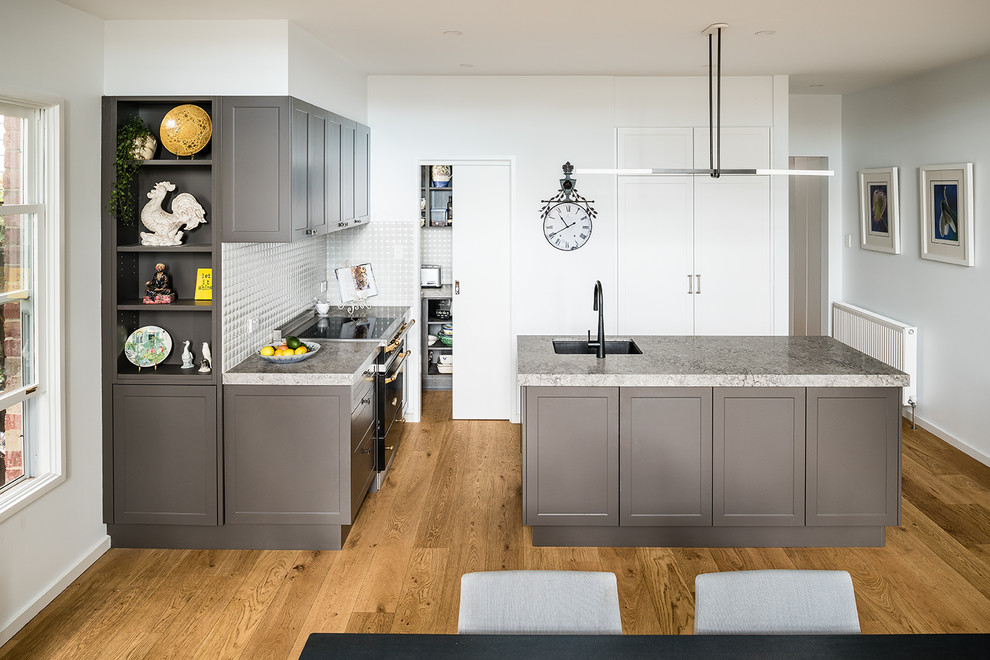 Design ideas for a small transitional l-shaped kitchen pantry in Melbourne with an undermount sink, shaker cabinets, grey cabinets, quartz benchtops, white splashback, ceramic splashback, black appliances, medium hardwood floors, with island and grey benchtop.