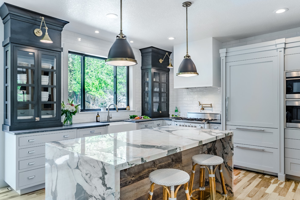 Mid-sized transitional kitchen in Denver with a farmhouse sink, marble benchtops, white splashback, ceramic splashback, panelled appliances, with island, glass-front cabinets, light hardwood floors, beige floor and black cabinets.