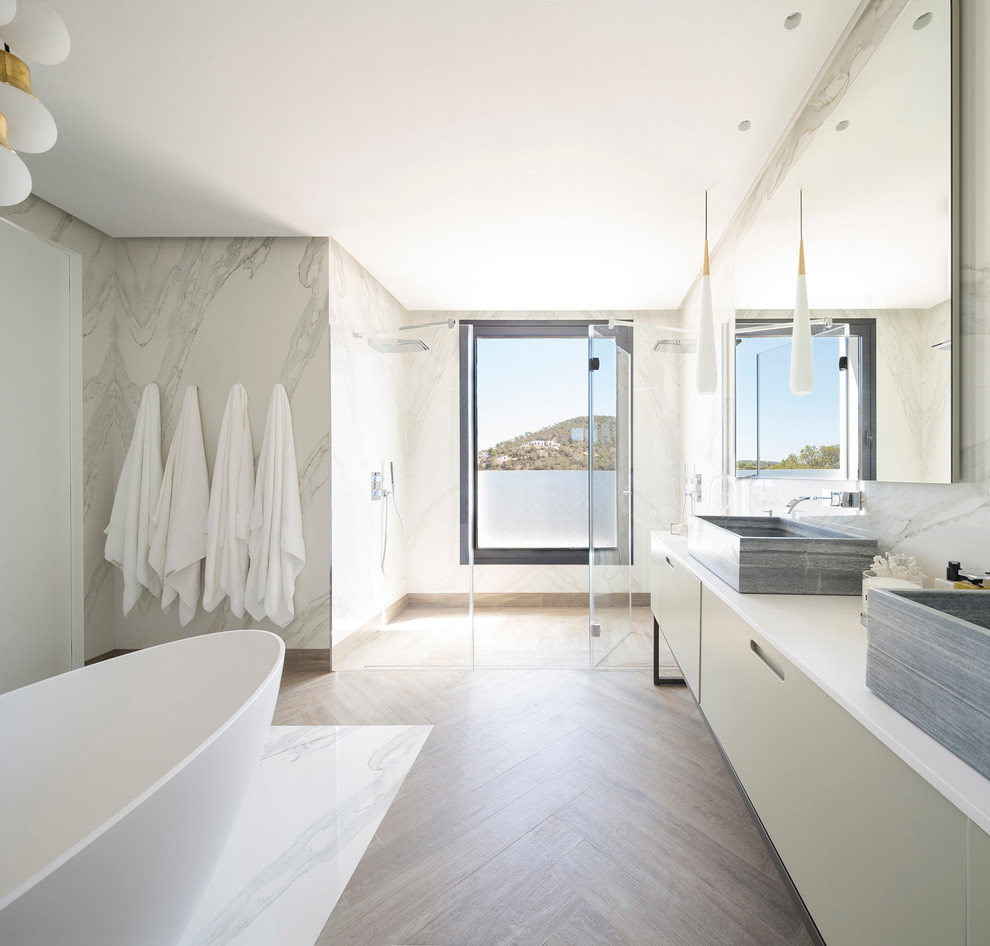 This is an example of an expansive contemporary master bathroom in Bilbao with flat-panel cabinets, white cabinets, a freestanding tub, a double shower, white walls, medium hardwood floors, a vessel sink and a hinged shower door.