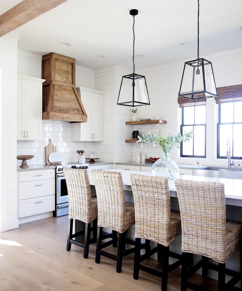 Photo of a country l-shaped kitchen in Other with a farmhouse sink, shaker cabinets, white cabinets, white splashback, stainless steel appliances, light hardwood floors, with island, beige floor and white benchtop.