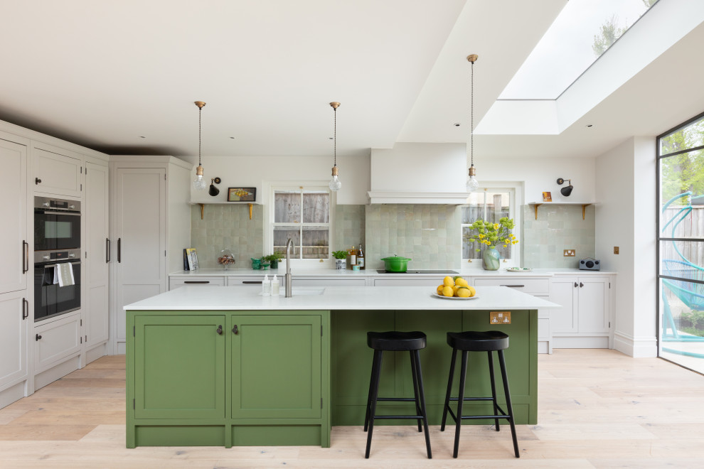 Design ideas for a medium sized classic l-shaped open plan kitchen in London with a submerged sink, shaker cabinets, green cabinets, composite countertops, green splashback, ceramic splashback, integrated appliances, light hardwood flooring, an island, white worktops and a vaulted ceiling.