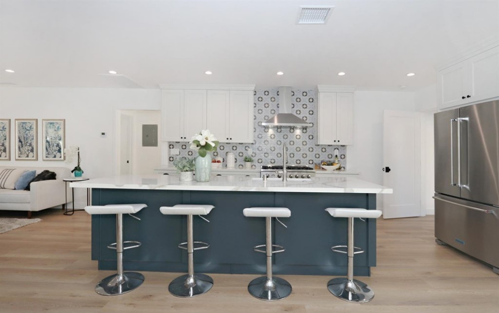 Photo of a mediterranean l-shaped kitchen in Los Angeles with a submerged sink, shaker cabinets, white cabinets, engineered stone countertops, multi-coloured splashback, ceramic splashback, stainless steel appliances, light hardwood flooring, an island, brown floors and white worktops.