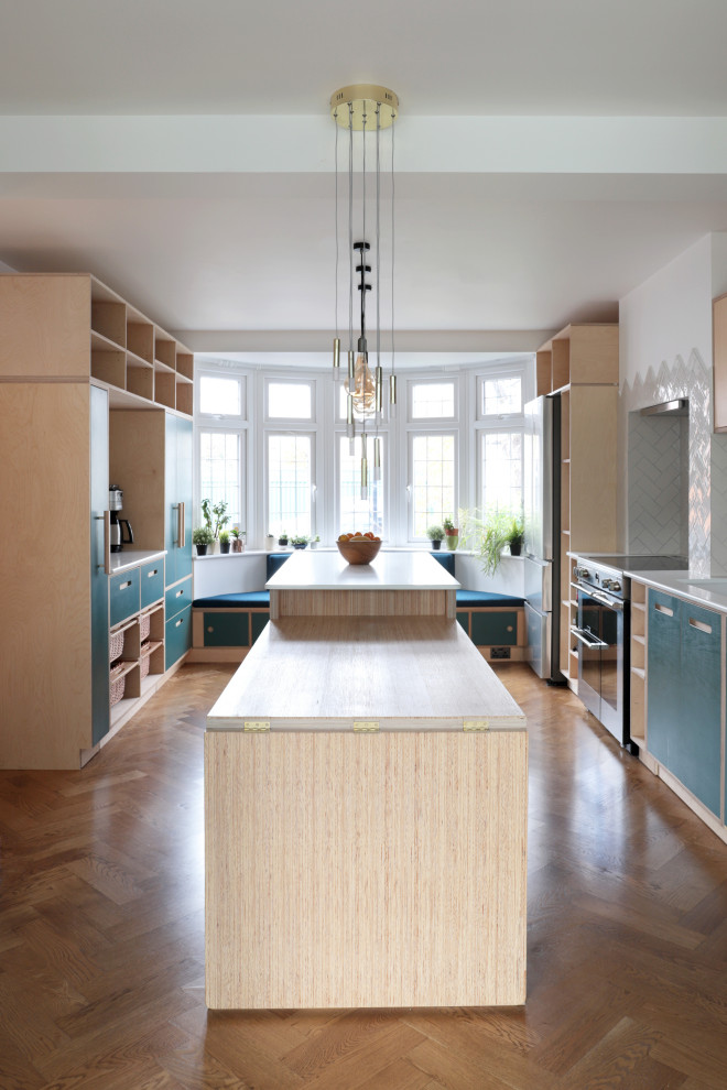 This is an example of a large contemporary galley open plan kitchen in London with a double-bowl sink, flat-panel cabinets, turquoise cabinets, white splashback, ceramic splashback, stainless steel appliances, medium hardwood floors, with island, brown floor and white benchtop.