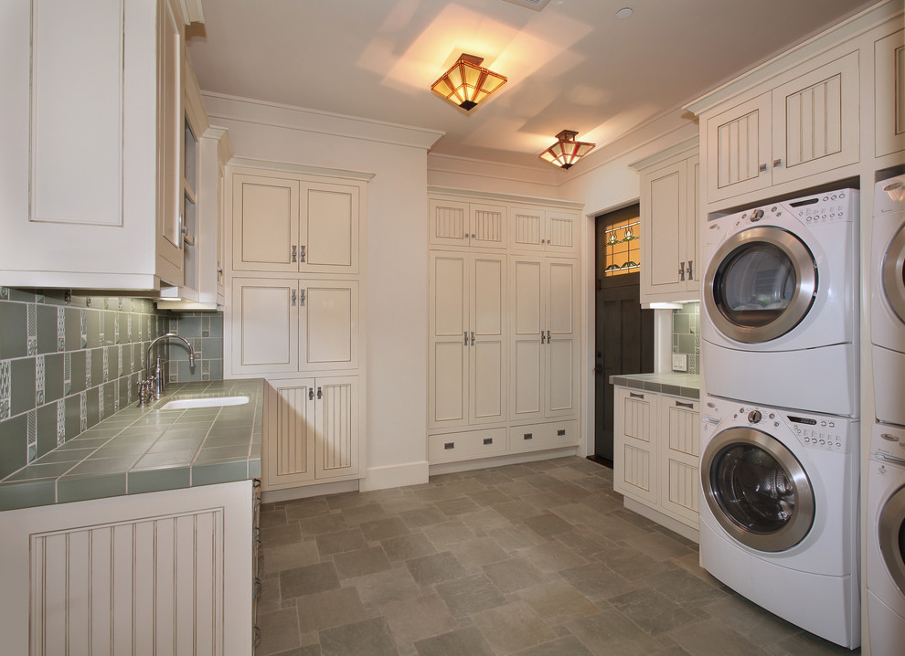 Photo of an expansive arts and crafts u-shaped dedicated laundry room in San Diego with recessed-panel cabinets, white walls, a stacked washer and dryer, grey floor, an undermount sink, tile benchtops, slate floors, green benchtop and beige cabinets.