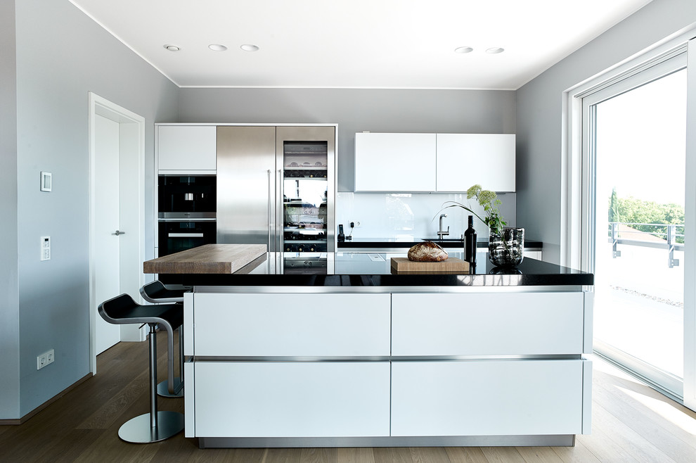 Mid-sized contemporary galley kitchen in Frankfurt with flat-panel cabinets, white cabinets, white splashback, glass sheet splashback, with island, black benchtop, an undermount sink, stainless steel appliances, medium hardwood floors and brown floor.