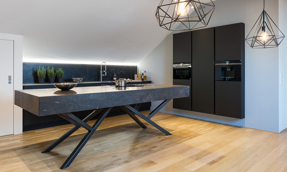 Design ideas for a large industrial l-shaped open plan kitchen in Munich with an undermount sink, flat-panel cabinets, black cabinets, granite benchtops, black splashback, stone slab splashback, black appliances, light hardwood floors, with island and brown floor.