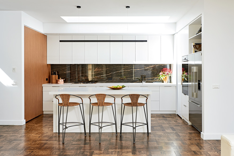 Photo of a mid-sized contemporary l-shaped kitchen in Melbourne with white cabinets, black splashback, stone slab splashback, stainless steel appliances, with island, brown floor, white benchtop, flat-panel cabinets and dark hardwood floors.