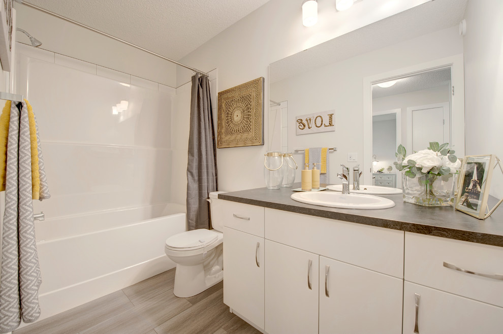 Mid-sized transitional kids bathroom in Edmonton with flat-panel cabinets, white cabinets, an alcove tub, a shower/bathtub combo, a two-piece toilet, white tile, ceramic tile, white walls, vinyl floors, a drop-in sink, laminate benchtops, grey floor, a shower curtain and black benchtops.