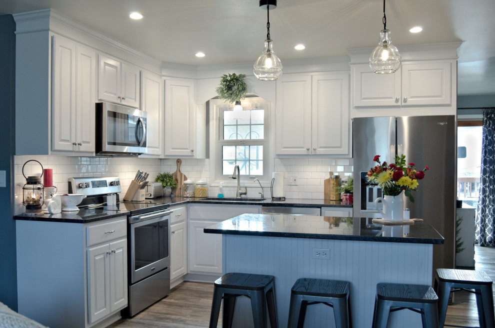 L-shaped kitchen photo with raised-panel cabinets, white cabinets, granite countertops, white backsplash, stainless steel appliances, an island and black countertops