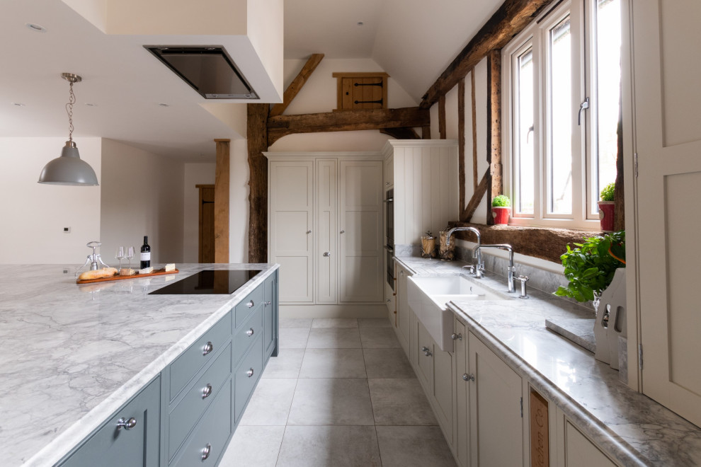 Photo of an expansive traditional cream and black single-wall open plan kitchen in Essex with a belfast sink, shaker cabinets, medium wood cabinets, quartz worktops, white splashback, engineered quartz splashback, black appliances, ceramic flooring, an island, grey floors, white worktops, exposed beams and a feature wall.