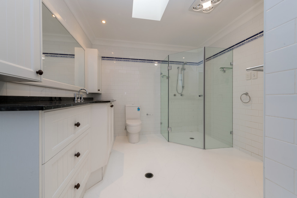 This is an example of a small traditional sauna bathroom in Sydney with white cabinets, a corner shower, a two-piece toilet, solid surface worktops, a hinged door, a single sink, a freestanding vanity unit, recessed-panel cabinets, a corner bath, white tiles, white walls, ceramic flooring, a submerged sink, white floors and black worktops.