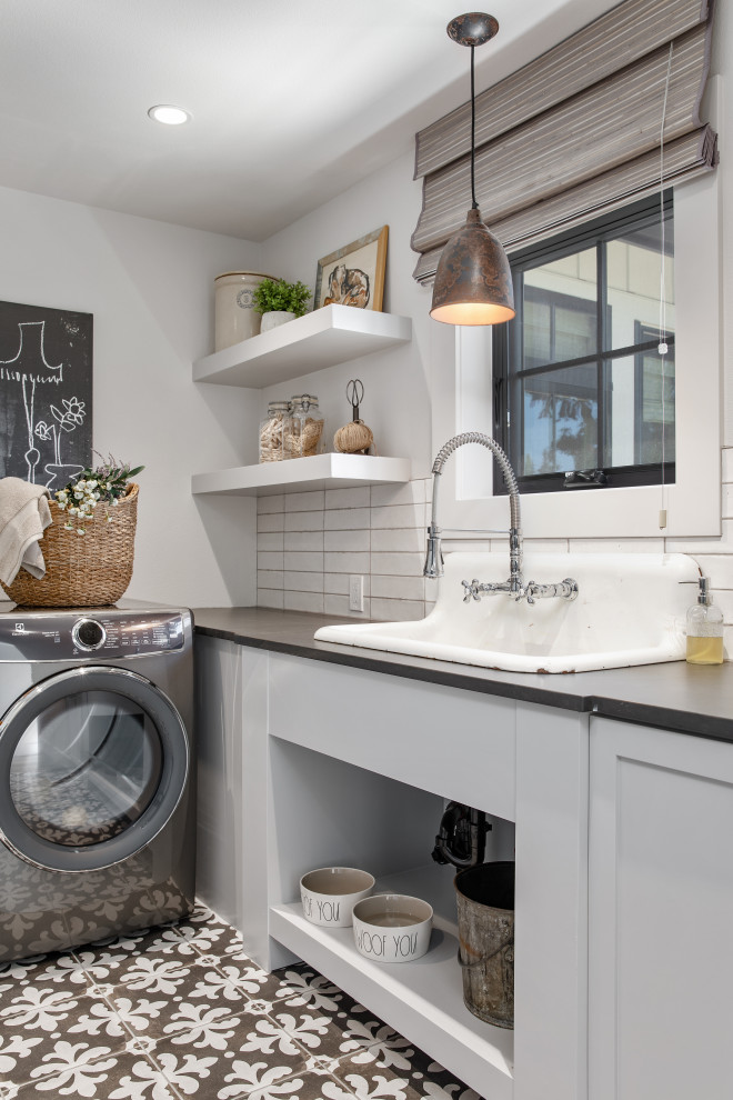 Photo of a mid-sized country single-wall dedicated laundry room in Portland with a farmhouse sink, shaker cabinets, grey cabinets, quartz benchtops, white splashback, ceramic splashback, white walls, ceramic floors, a side-by-side washer and dryer, black floor and black benchtop.