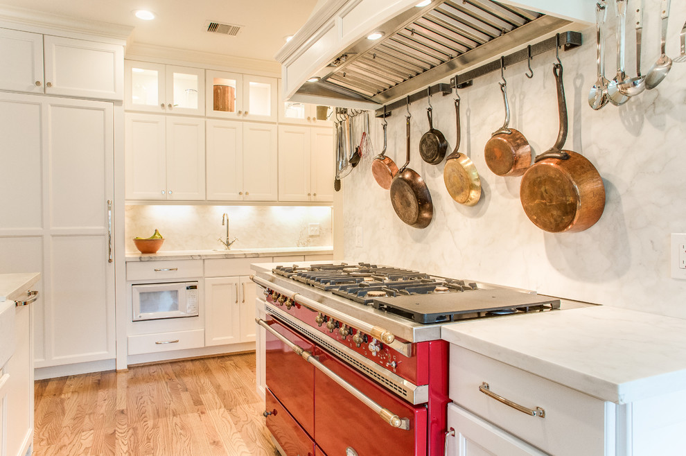 Mid-sized transitional l-shaped separate kitchen in Houston with a farmhouse sink, recessed-panel cabinets, white cabinets, marble benchtops, white splashback, stone slab splashback, panelled appliances, light hardwood floors and with island.