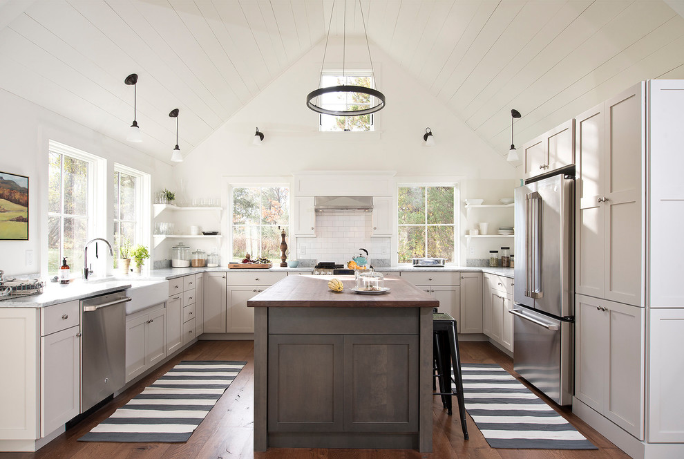 Country u-shaped kitchen in New York with a farmhouse sink, shaker cabinets, grey cabinets, wood benchtops, white splashback, subway tile splashback, stainless steel appliances, medium hardwood floors, with island and brown floor.
