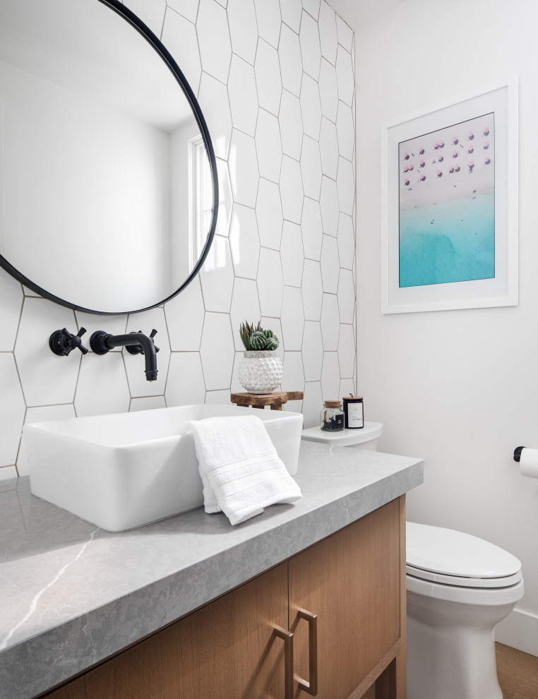 Mid-sized beach style powder room in Orange County with flat-panel cabinets, brown cabinets, a two-piece toilet, white tile, ceramic tile, white walls, light hardwood floors, a vessel sink, engineered quartz benchtops, beige floor and grey benchtops.