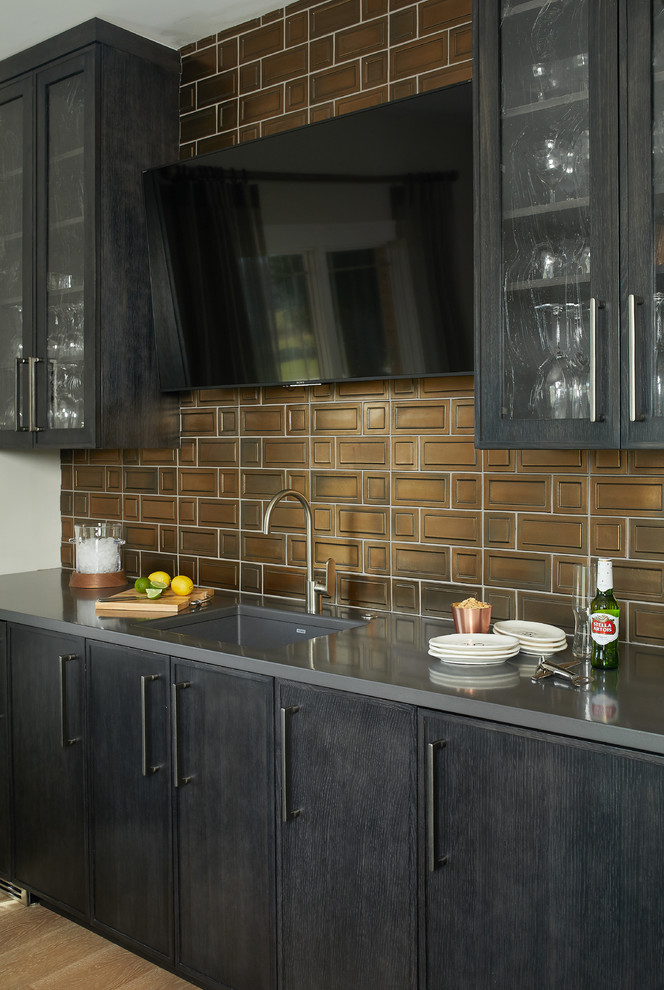 This is an example of a mid-sized transitional single-wall wet bar in Grand Rapids with an undermount sink, black cabinets, solid surface benchtops, brown splashback, light hardwood floors, brown floor, glass-front cabinets, metal splashback and grey benchtop.
