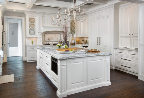 Luxury white kitchen cabinet design with a large marble island, glass-door uppers and built-in appliances on dark wood flooring.