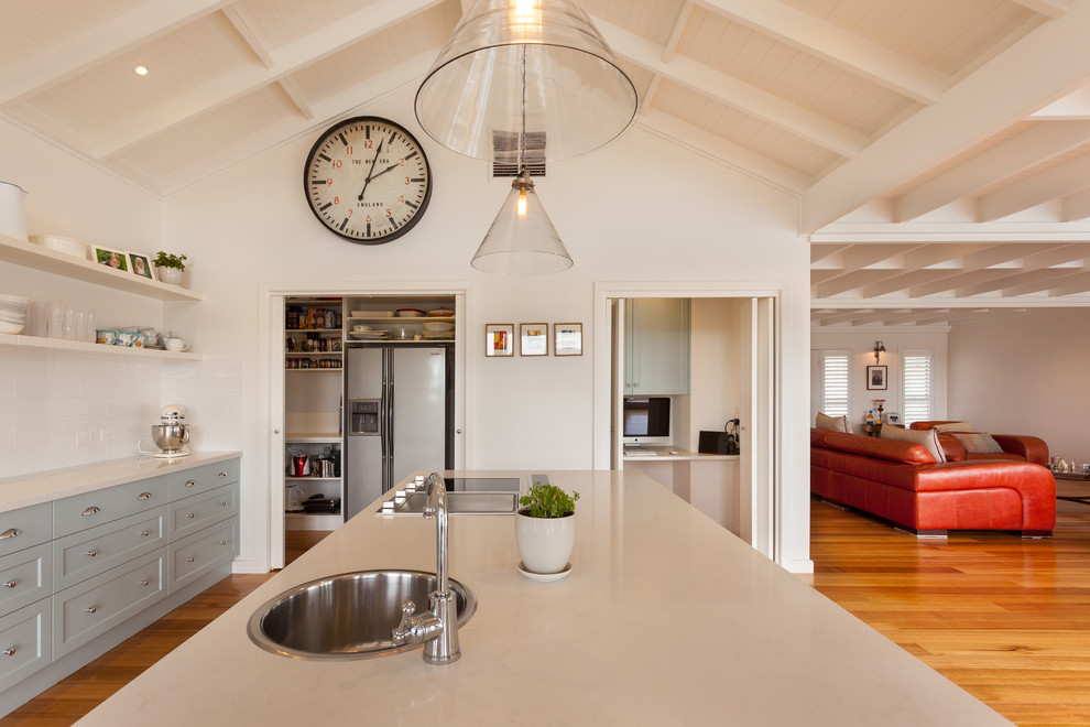 This is an example of a mid-sized beach style galley open plan kitchen in Other with a drop-in sink, shaker cabinets, blue cabinets, quartz benchtops, white splashback, ceramic splashback, stainless steel appliances, medium hardwood floors, with island, brown floor and grey benchtop.