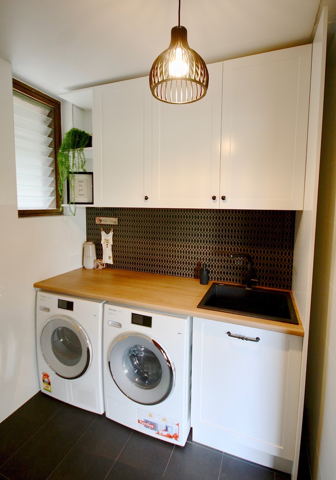 Photo of a small traditional single-wall dedicated laundry room in Sydney with a drop-in sink, shaker cabinets, white cabinets, wood benchtops, white walls, ceramic floors, a side-by-side washer and dryer and black floor.