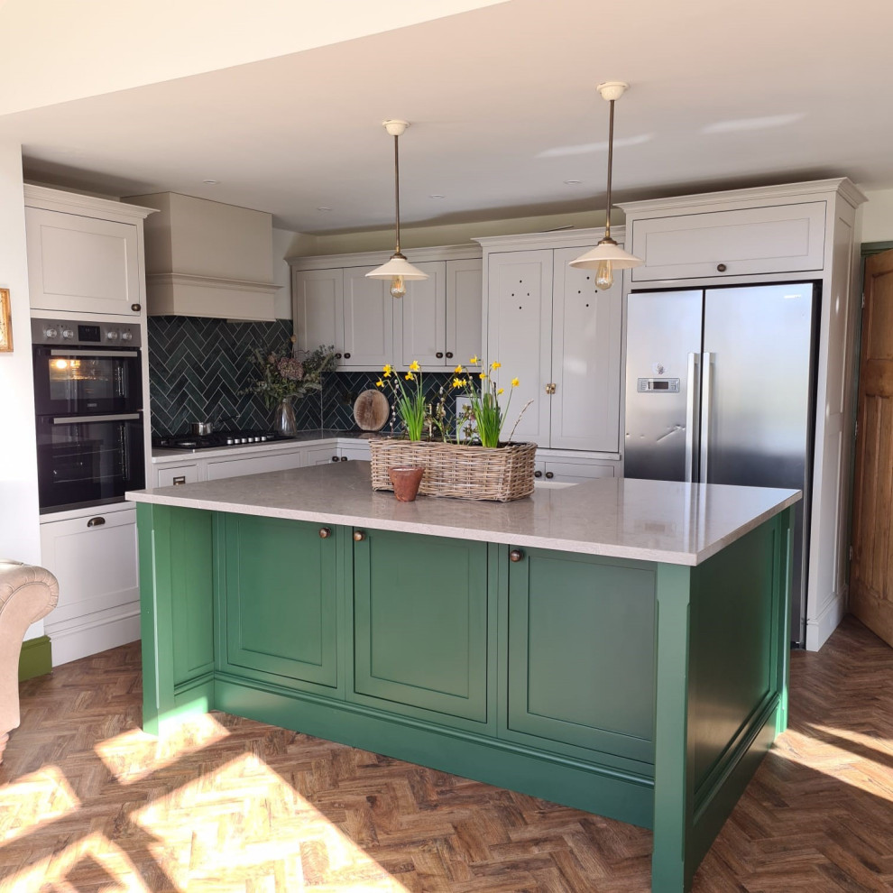 This is an example of a medium sized farmhouse l-shaped kitchen/diner in West Midlands with a belfast sink, beaded cabinets, grey cabinets, quartz worktops, green splashback, ceramic splashback, integrated appliances, laminate floors, an island, brown floors and white worktops.