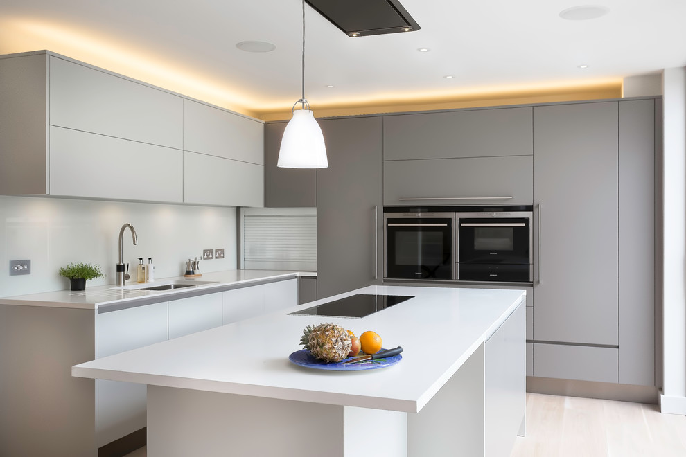 This is an example of a mid-sized contemporary l-shaped kitchen in Cambridgeshire with flat-panel cabinets, grey cabinets, solid surface benchtops, white splashback, glass sheet splashback, stainless steel appliances, limestone floors and with island.