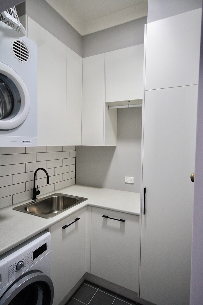 This is an example of a small transitional l-shaped dedicated laundry room in Sydney with a drop-in sink, flat-panel cabinets, white cabinets, laminate benchtops, white walls, ceramic floors, a stacked washer and dryer and grey floor.