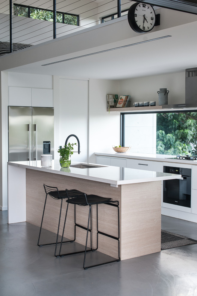 This is an example of a contemporary l-shaped kitchen in Gold Coast - Tweed with a single-bowl sink, flat-panel cabinets, white cabinets, window splashback, stainless steel appliances, concrete floors, with island, grey floor and white benchtop.