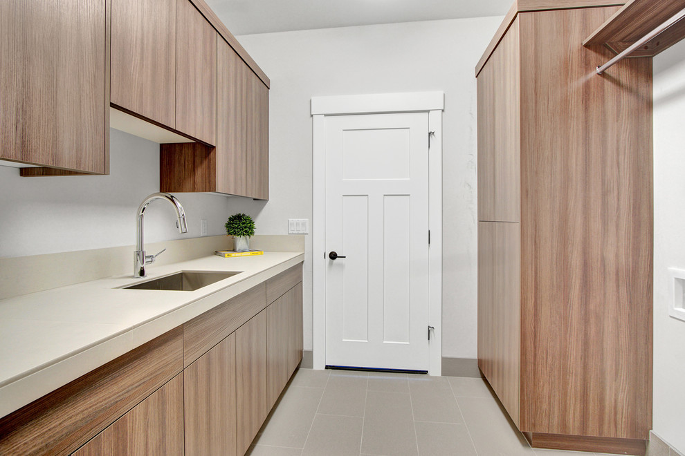 Photo of a small arts and crafts galley dedicated laundry room in Other with a drop-in sink, flat-panel cabinets, tile benchtops, grey walls, ceramic floors, a side-by-side washer and dryer, grey floor and medium wood cabinets.