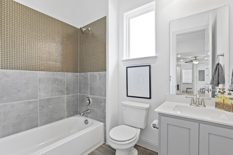 Photo of a mid-sized contemporary kids bathroom in Dallas with recessed-panel cabinets, grey cabinets, an alcove tub, a shower/bathtub combo, ceramic tile, white walls, medium hardwood floors, an undermount sink, solid surface benchtops, beige floor, white benchtops and multi-coloured tile.