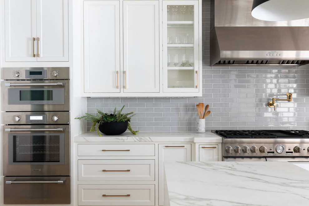 Mid-sized transitional l-shaped kitchen pantry in Sacramento with a farmhouse sink, shaker cabinets, white cabinets, solid surface benchtops, grey splashback, ceramic splashback, stainless steel appliances, light hardwood floors, with island and white benchtop.