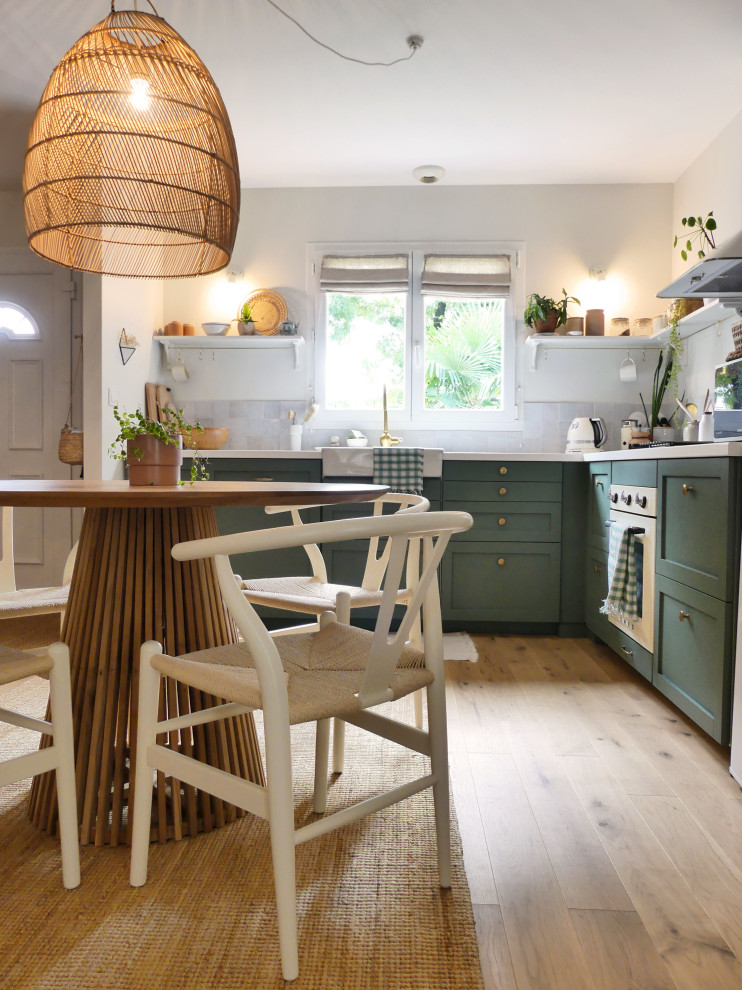 Inspiration for a mid-sized beach style l-shaped open plan kitchen in Nantes with a farmhouse sink, green cabinets, laminate benchtops, beige splashback, ceramic splashback, white appliances, light hardwood floors and white benchtop.
