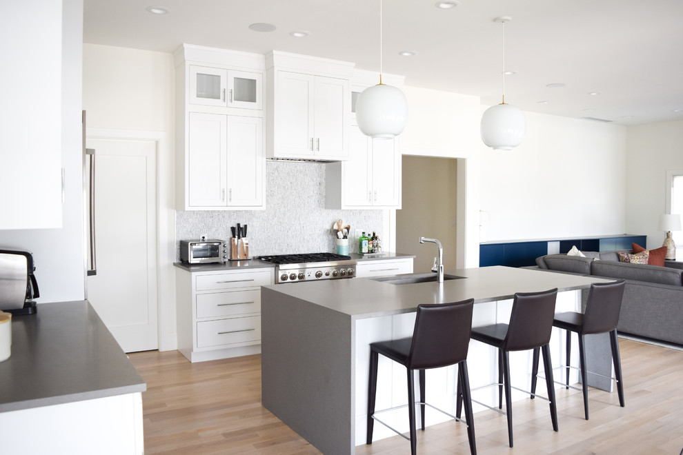 Design ideas for a mid-sized beach style l-shaped eat-in kitchen in New York with an undermount sink, recessed-panel cabinets, white cabinets, quartz benchtops, grey splashback, marble splashback, stainless steel appliances, light hardwood floors and with island.