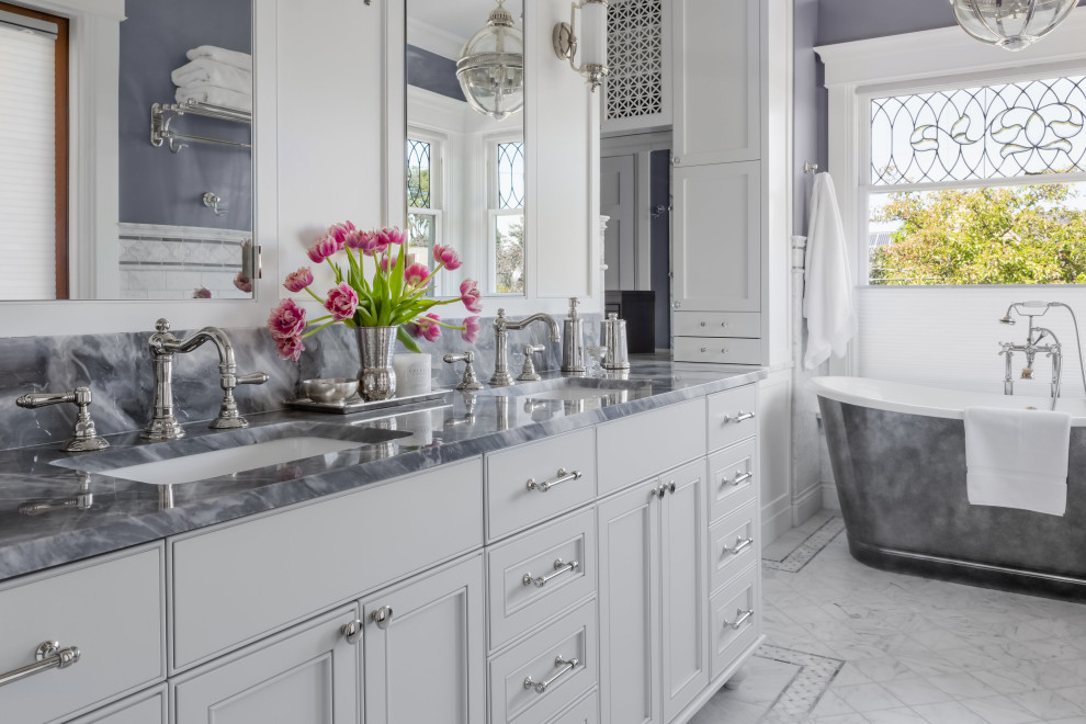 This is an example of a mid-sized arts and crafts master bathroom in Seattle with recessed-panel cabinets, grey cabinets, a freestanding tub, a double shower, a two-piece toilet, white tile, marble, grey walls, marble floors, an undermount sink, marble benchtops, white floor, a hinged shower door, grey benchtops, a shower seat, a double vanity and a freestanding vanity.