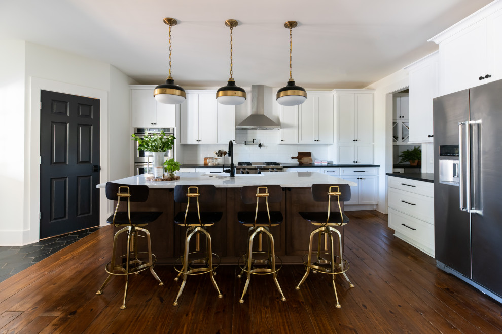 Design ideas for a large country l-shaped kitchen in Nashville with a farmhouse sink, shaker cabinets, white cabinets, white splashback, subway tile splashback, stainless steel appliances, medium hardwood floors, with island, brown floor and black benchtop.