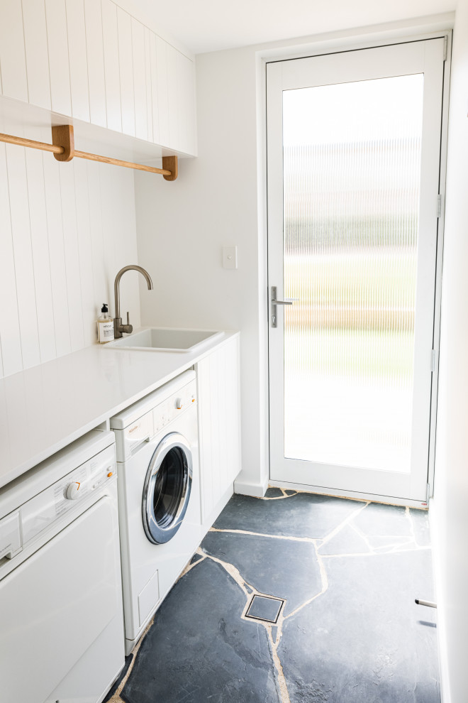 Photo of a medium sized beach style single-wall separated utility room in Adelaide with a built-in sink, shaker cabinets, white cabinets, laminate countertops, white splashback, tonge and groove splashback, white walls, slate flooring, an integrated washer and dryer, grey floors and white worktops.
