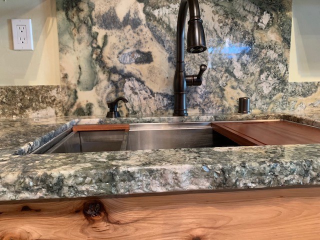 Example of a mid-sized mountain style medium tone wood floor and brown floor open concept kitchen design with an undermount sink, flat-panel cabinets, light wood cabinets, granite countertops, gray backsplash, stone slab backsplash, stainless steel appliances and gray countertops