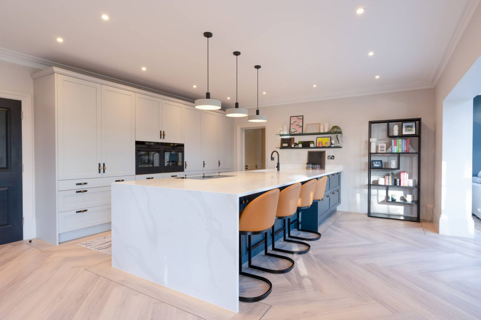 Large classic single-wall open plan kitchen in Hertfordshire with a single-bowl sink, recessed-panel cabinets, grey cabinets, quartz worktops, black appliances, light hardwood flooring, an island, brown floors and white worktops.