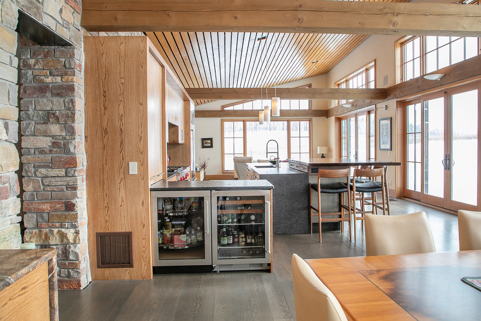Example of a large mountain style l-shaped medium tone wood floor and brown floor open concept kitchen design with flat-panel cabinets, light wood cabinets, granite countertops, black backsplash, stainless steel appliances, an island, black countertops, a double-bowl sink and porcelain backsplash