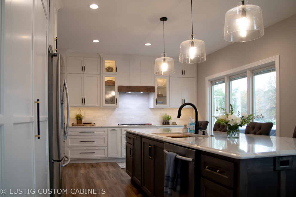 Open concept kitchen - large transitional l-shaped laminate floor and brown floor open concept kitchen idea with an undermount sink, flat-panel cabinets, white cabinets, quartz countertops, white backsplash, ceramic backsplash, stainless steel appliances, an island and white countertops