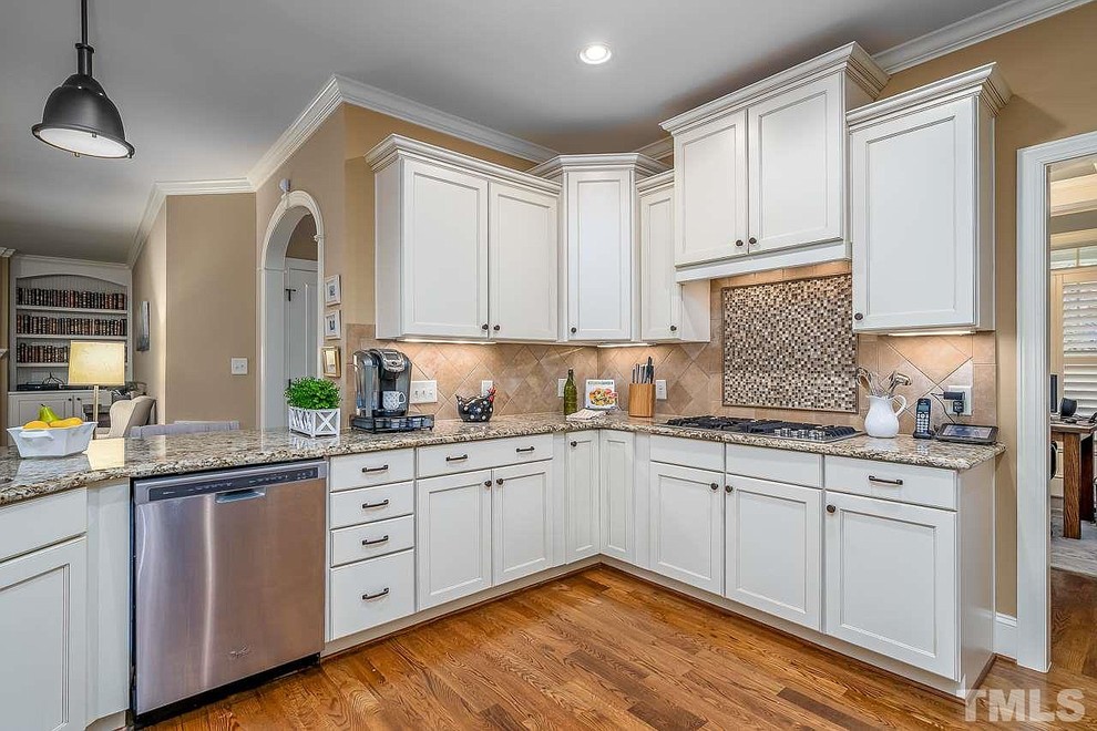 This is an example of a mid-sized transitional u-shaped kitchen in Raleigh with an undermount sink, recessed-panel cabinets, white cabinets, granite benchtops, beige splashback, travertine splashback, stainless steel appliances, medium hardwood floors, no island, brown floor and multi-coloured benchtop.