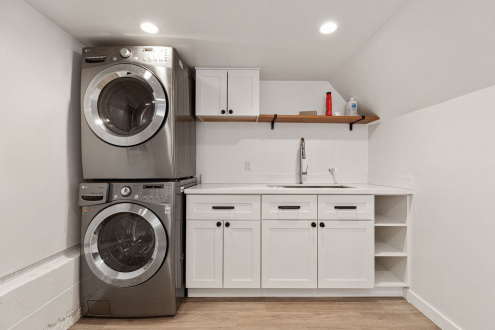 Photo of a small traditional single-wall dedicated laundry room in San Francisco with an undermount sink, shaker cabinets, white cabinets, quartz benchtops, white splashback, engineered quartz splashback, white walls, light hardwood floors, a stacked washer and dryer, white floor and white benchtop.