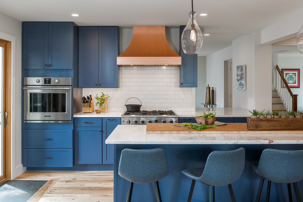 This is an example of a large transitional open plan kitchen in Burlington with shaker cabinets, blue cabinets, quartzite benchtops, white splashback, subway tile splashback, stainless steel appliances, light hardwood floors, with island, white benchtop and beige floor.