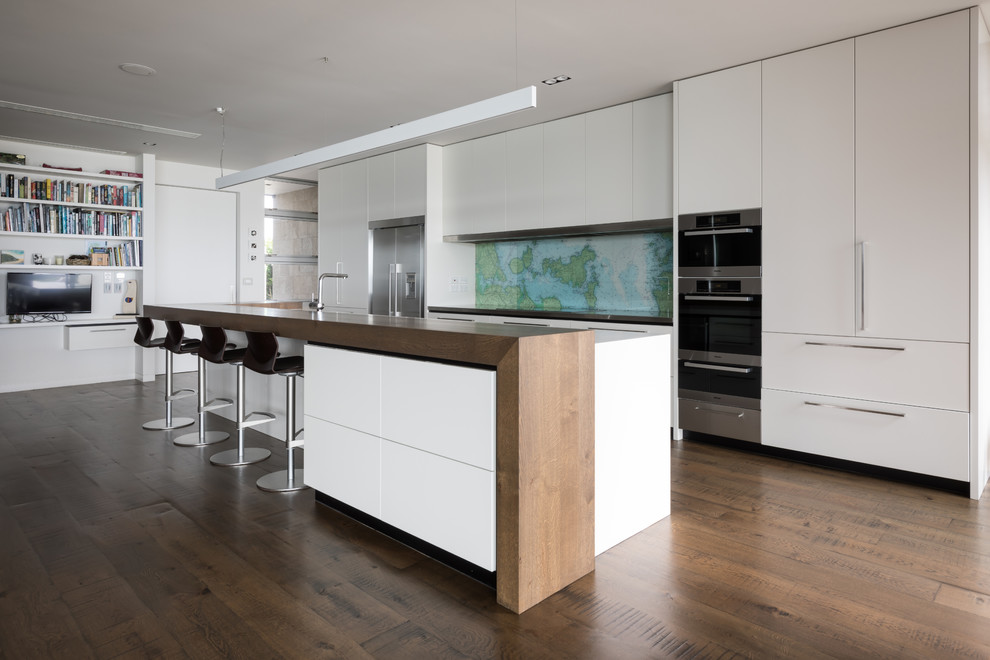 Contemporary galley kitchen in Auckland with flat-panel cabinets, white cabinets, wood benchtops, multi-coloured splashback, stainless steel appliances, medium hardwood floors, with island, brown floor and brown benchtop.