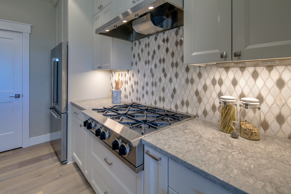 Example of a mid-sized arts and crafts l-shaped medium tone wood floor and brown floor open concept kitchen design with an undermount sink, recessed-panel cabinets, gray cabinets, quartz countertops, white backsplash, ceramic backsplash, stainless steel appliances, an island and gray countertops