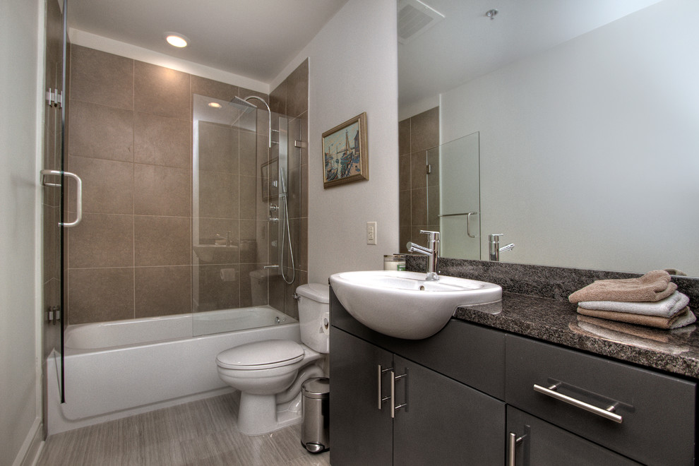 Design ideas for a mid-sized transitional 3/4 bathroom in Miami with flat-panel cabinets, black cabinets, an alcove tub, a shower/bathtub combo, a two-piece toilet, white walls, porcelain floors, a drop-in sink, granite benchtops, grey floor, a hinged shower door and multi-coloured benchtops.
