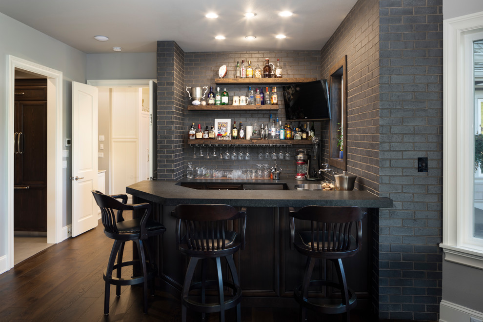 This is an example of a mid-sized transitional galley seated home bar in Portland with a drop-in sink, recessed-panel cabinets, dark wood cabinets, granite benchtops, grey splashback, brick splashback, medium hardwood floors, brown floor and black benchtop.