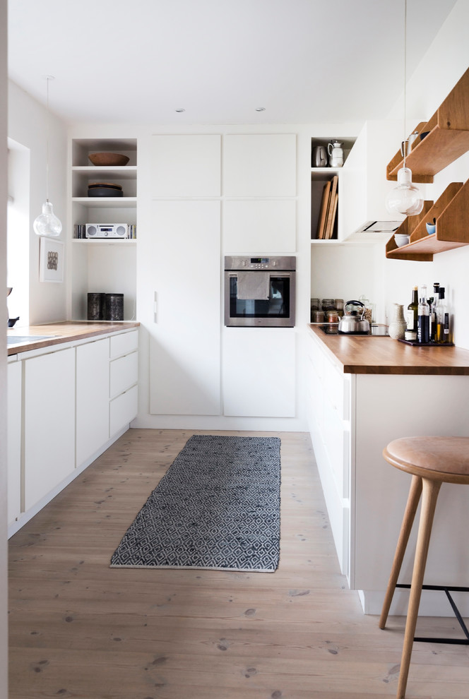 Inspiration for a mid-sized scandinavian galley separate kitchen in Odense with flat-panel cabinets, white cabinets, wood benchtops, panelled appliances, light hardwood floors, beige floor, a drop-in sink, white splashback, no island and brown benchtop.