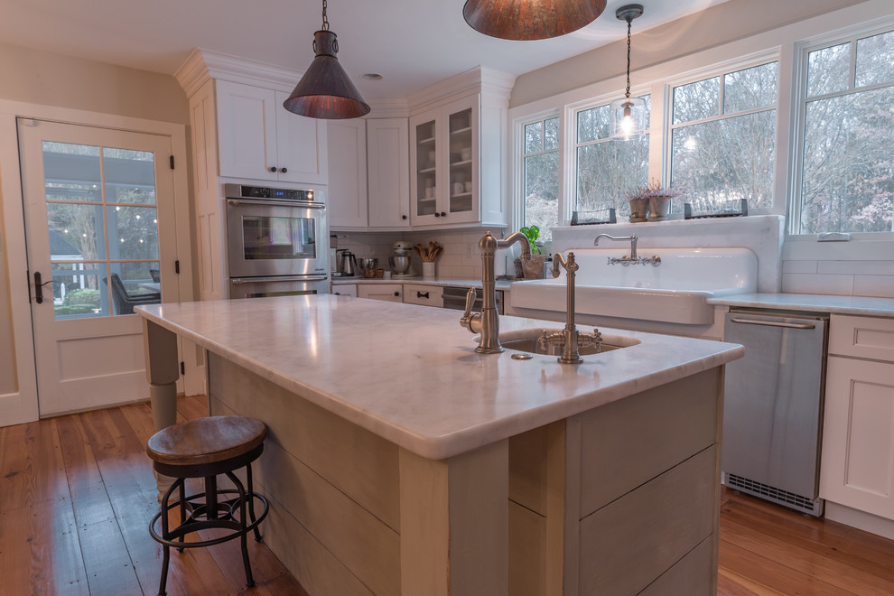 Kitchen - mid-sized transitional medium tone wood floor kitchen idea with a farmhouse sink, recessed-panel cabinets, white cabinets, marble countertops, white backsplash, subway tile backsplash, stainless steel appliances and an island
