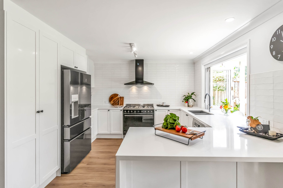 Photo of a mid-sized transitional u-shaped open plan kitchen in Sydney with an undermount sink, shaker cabinets, white cabinets, quartz benchtops, white splashback, subway tile splashback, stainless steel appliances, vinyl floors, a peninsula, brown floor and white benchtop.